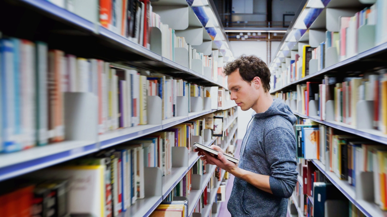 Junger Mann steht zwischen Regalen in einer Bibliothek und schaut in ein Buch.