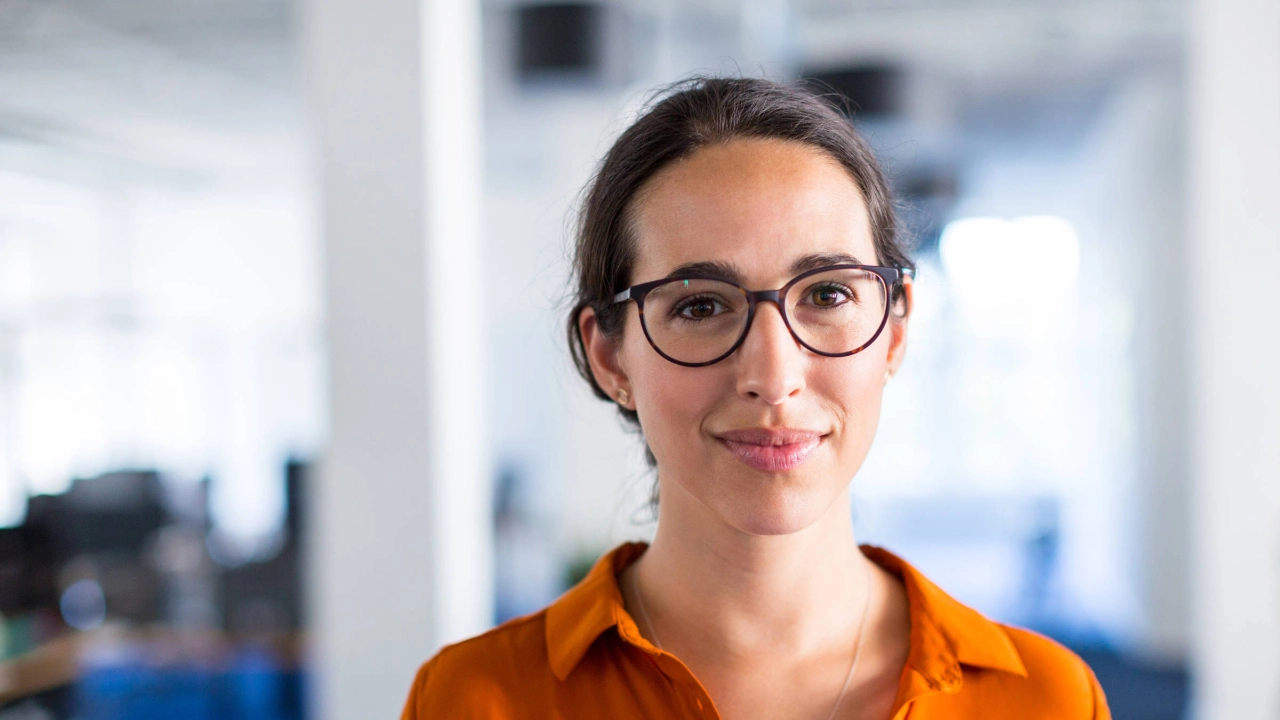 Eine junge Frau mit oranger Bluse und Brille lächelt in die Kamera.
