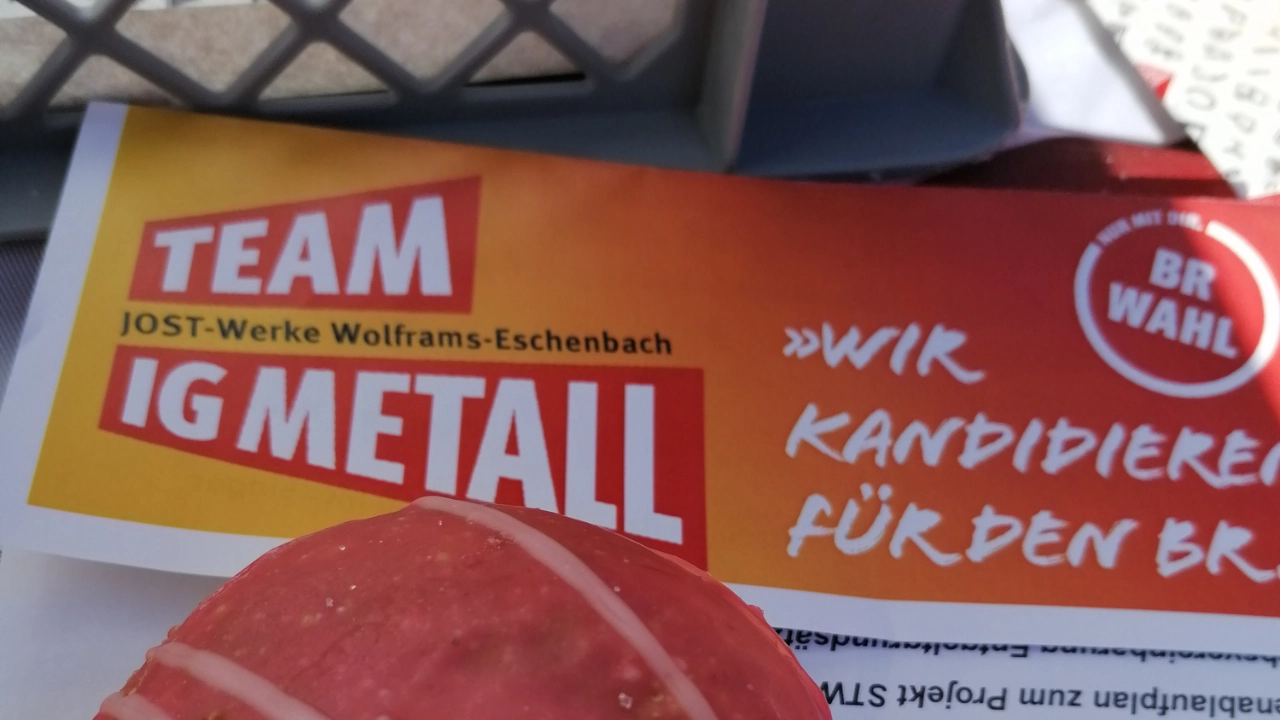 Ein rot-weißer Krapfen liegt gemeinsam mit einem Wahlflyer für die Kandidat*innen der IG Metall-Liste zur BR-Wahl 2022 und einer IG Metall-Serviette auf einem Tisch.