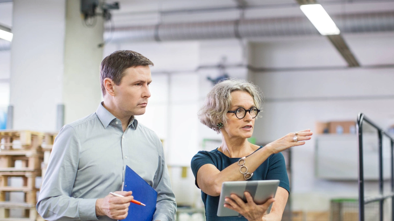 Eine Frau und ein Mann stehengestikulierend in einer Produktionsstätte und unterhalten sich.
