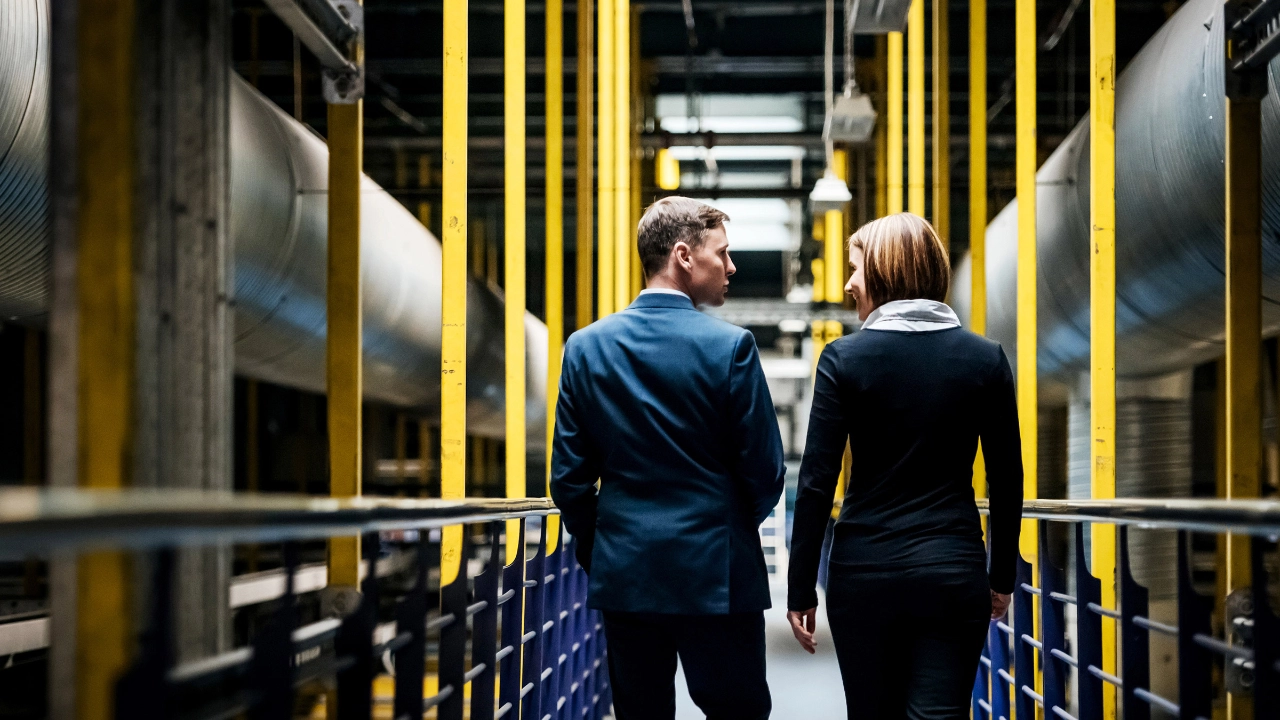 Eine Frau und ein Mann gehen durch eine Frabrik und unterhalten sich dabei.