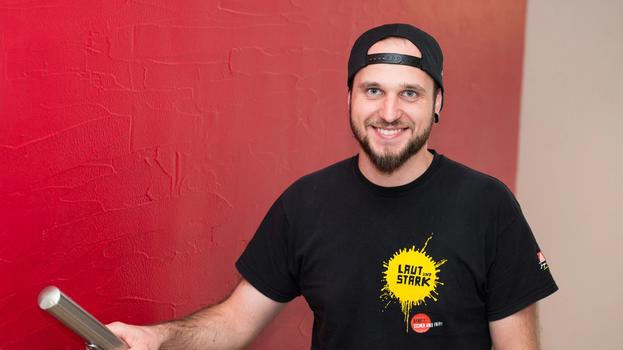 Junger Mann mit IG Metall-Kampagnen-Shirt blickt lächelnd direkt in die Kamera.