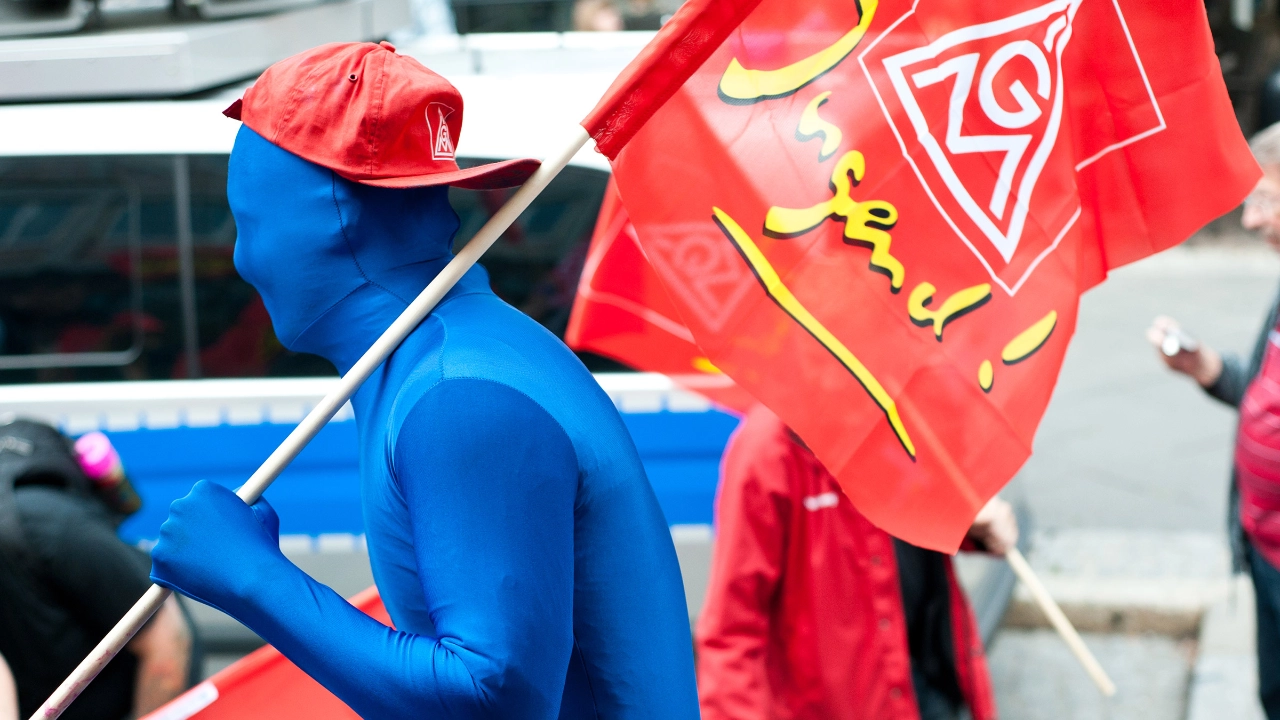 Junger Erwachsener mit blauem Morphsuit, IG Metall-Kappe und IG Metall Jugend-Fahne auf Demo.