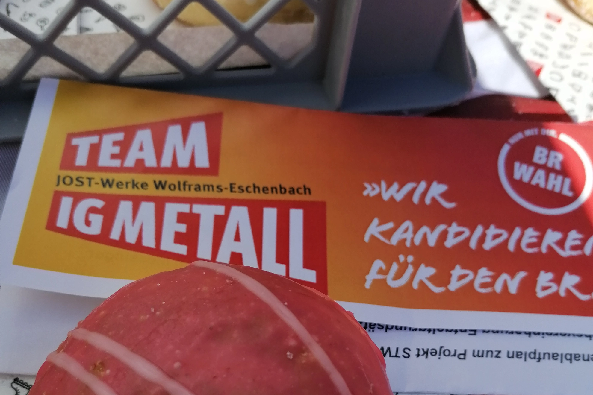 Ein rot-weißer Krapfen liegt gemeinsam mit einem Wahlflyer für die Kandidat*innen der IG Metall-Liste zur BR-Wahl 2022 und einer IG Metall-Serviette auf einem Tisch.